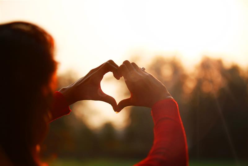 Takaa päin otettu lähikuva ihmisestä, joka muodostaa sormillaan sydämen. Taustalla aurinkoinen maisema.