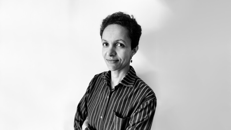 Raidalliseen kauluspaitaan pukeutunut Tummahiuksinen nainen katsoo kädet rinnalle ristittynä hymyillen kameraan.