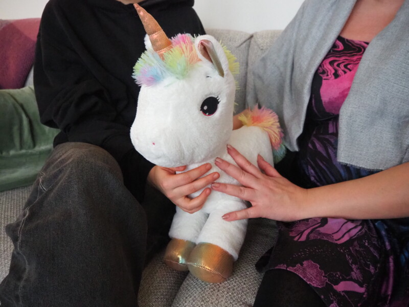 Kaksi ihmistä istuu rinnakkain sohvalla, heillä on välissään yksisarvispehmolelu.