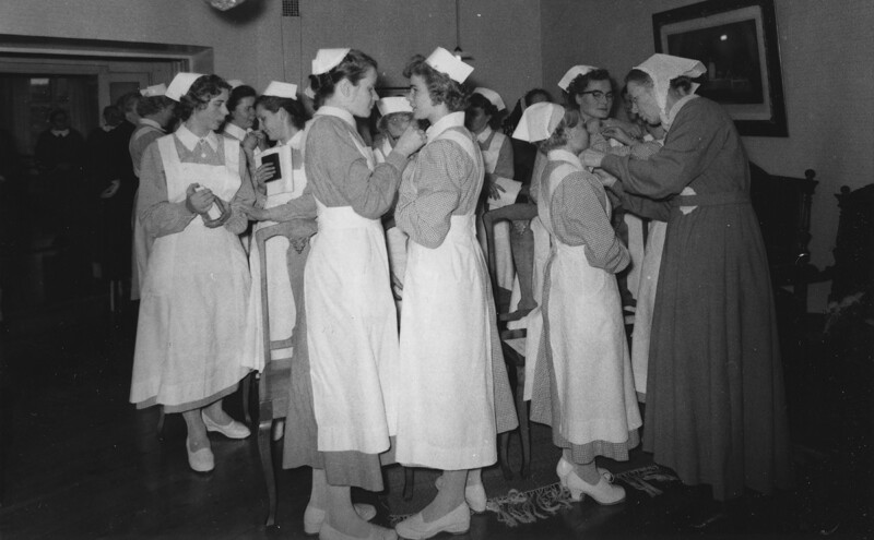 Mustavalkoinen kuva ryhmästä naispuolisia apuhoitajia, jotka ovat rivissä sisätiloissa, osa heistä keskustelee.