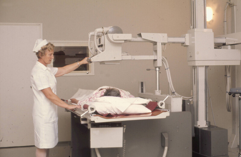 Sairaanhoitaja säätää röntgenlaitetta potilaan ollessa makuulla tutkimuspöydällä sairaalaympäristössä.