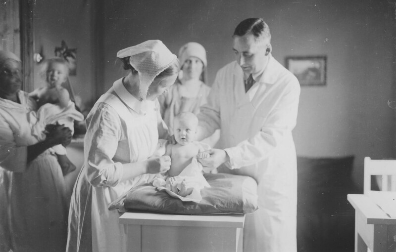 Pieni lapsi istuu hoitopöydällä. Häntä tutkivat lääkäri ja diakonissa.