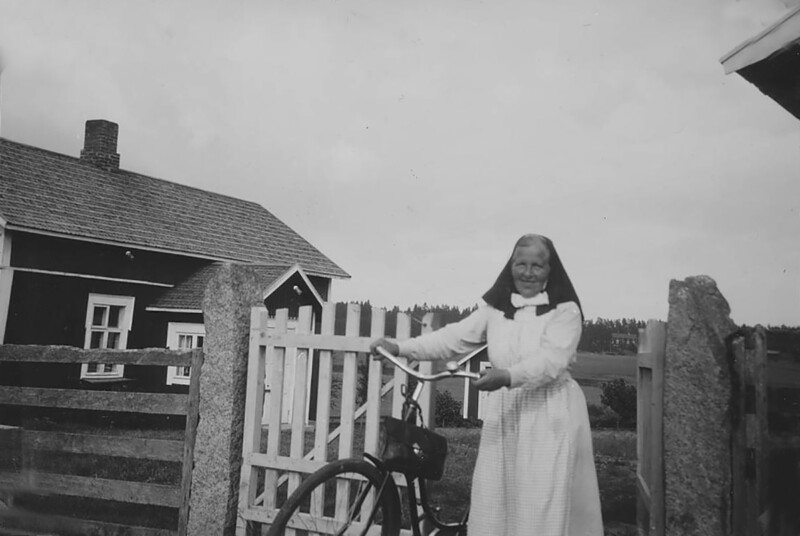 Mustavalkoinen kuva, jossa diakonissa seisoo polkupyörän kanssa maatilan pihalla, taustalla talo ja aita.