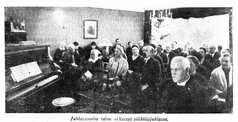 Mustavalkoinen valokuva juhlatilaisuudesta, jossa suuri joukko ihmisiä istuu tiiviisti yhdessä huoneessa, taustalla piano ja seinällä taulu.