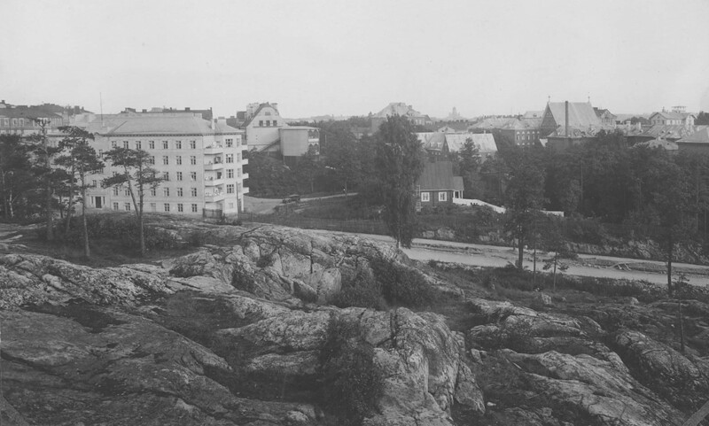 Mustavalkoinen valokuva, jossa näkyy kallioinen etualue ja taustalla useita rakennuksia sekä puita, kuva esittää Elim-rakennusta ja sen ympäristöä Diakonissalaitoksen korttelissa.