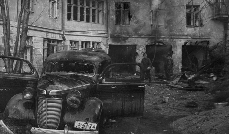 Musta-valkoinen kuva pommituksen jäljiltä, jossa tuhoutunut auto ja vaurioitunut rakennus taustalla.