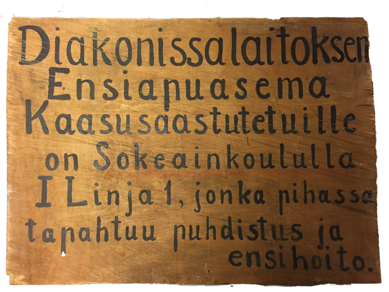 Käsinmaalattu puisessa kyltissä teksti: "Diakonissalaitoksen Ensiapuasema Kaasusaastutetuille on Sokeainkoululla II linja 1, jonka pihassa tapahtuu puhdistus ja ensihoito."