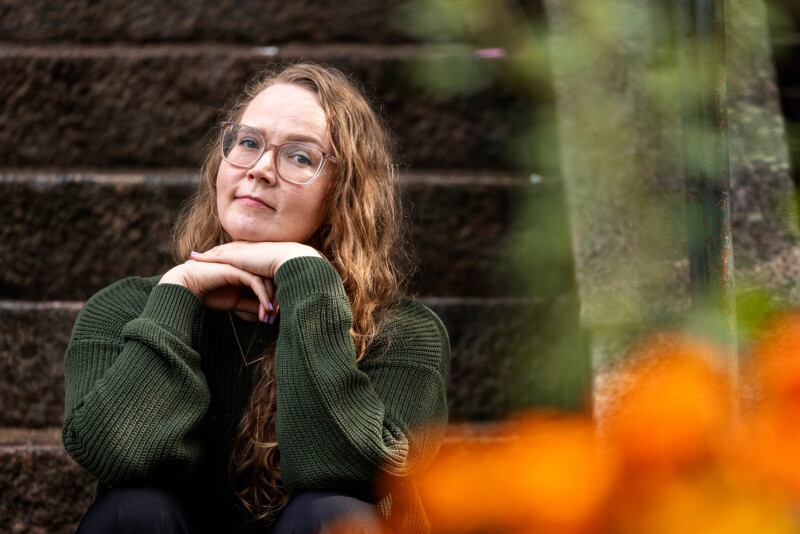 Nuorehko nainen vihreässä neuleessa istuu ja nojaa käsiinsä, taustalla kiviportaikko ja etualalla sumeita oransseja kukkia.