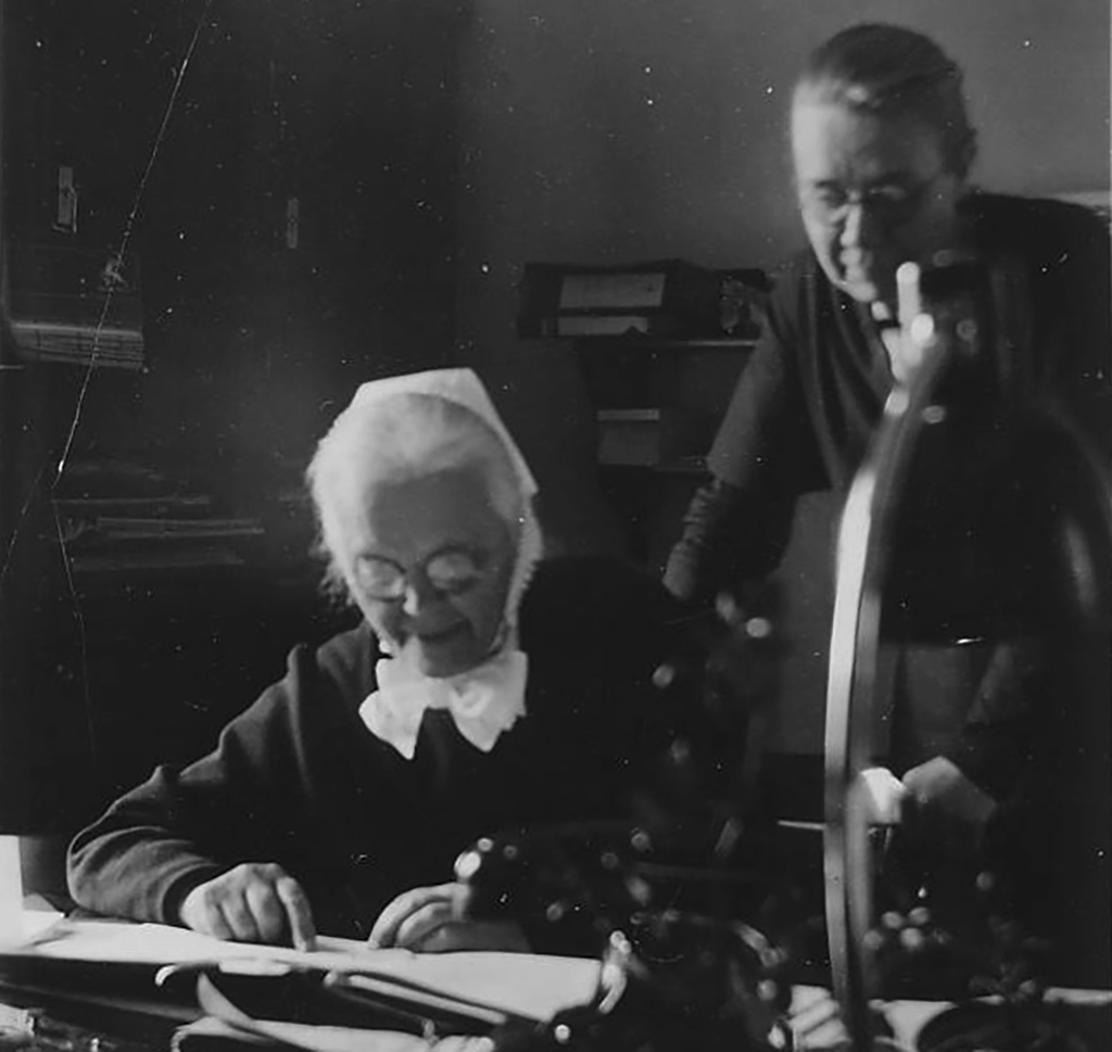 Mustavalkoinen valokuva, jossa vanhempi nainen valkopäähineessä ja lasit päässään istuu pöydän ääressä lukemassa tai kirjoittamassa, ja toinen henkilö nojaa pöytään hänen takanaan katsellen hänen tekemisiään.