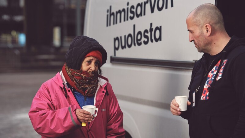 Punatakkinen nainen seisoo auton vieressä. Hän keskustelee miehen kanssa. Auto on Hirundo Mobiili.