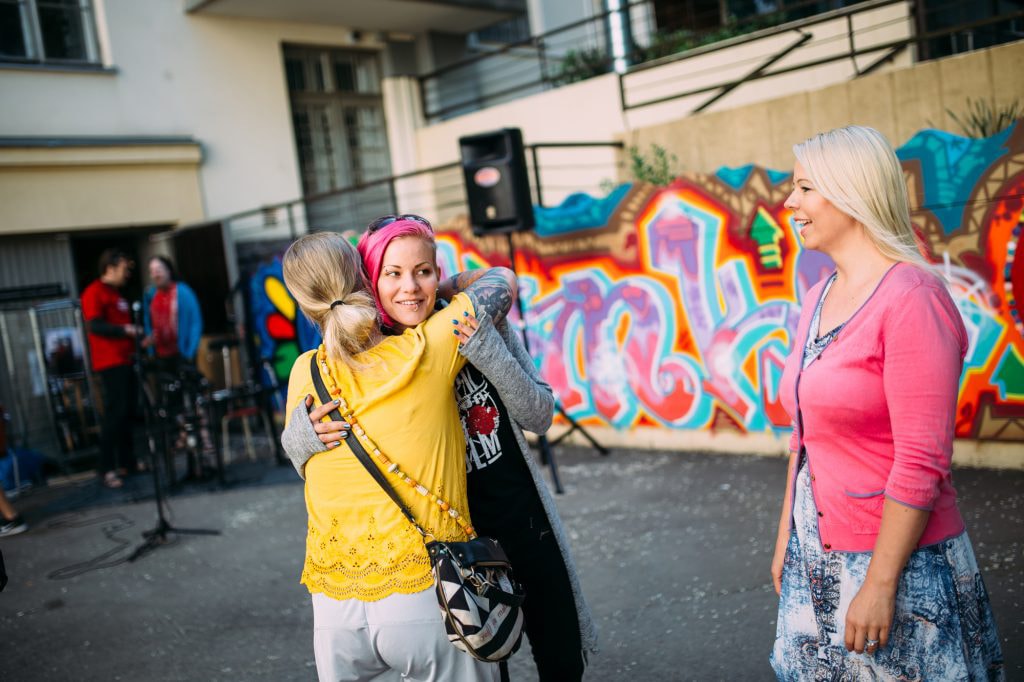 Kaksi naista halaa, kolmas seisoo vieressä ja nauraa.