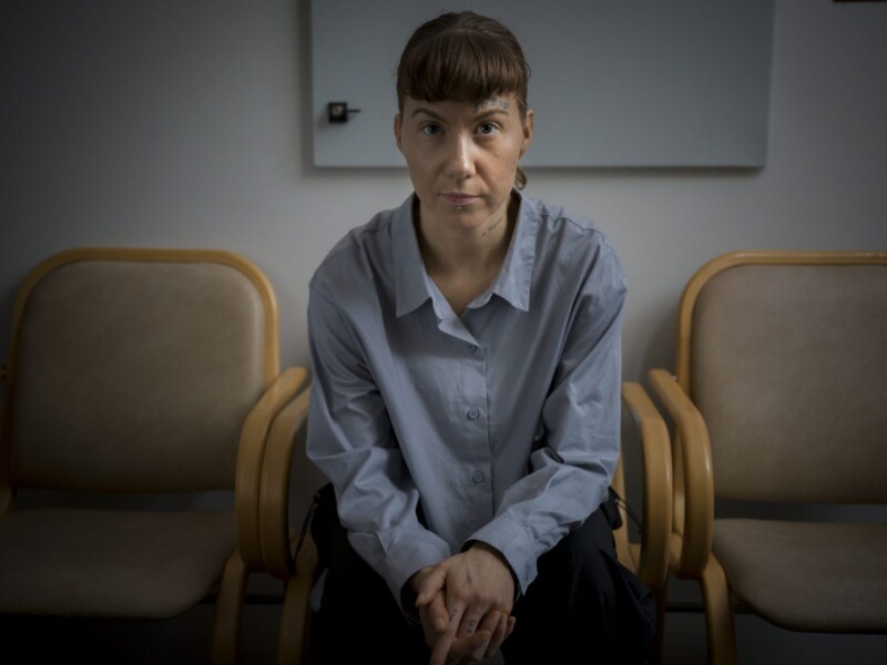 Vaaleansiniseen paitaan pukeutunut nuori nainen istuu tuolilla ja katsoo vakavana kameraan.