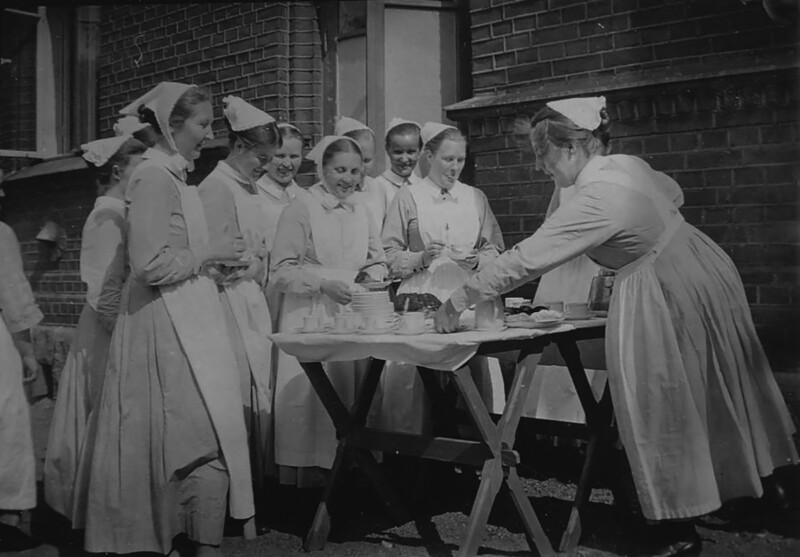Mustavalkoinen kuva, jossa joukko diakonissoja tai sairaanhoitajia pukeutuneina perinteisiin univormuihinsa, kokoontuneena ulkona pöydän ympärille nauttimaan kahvitaukoa.