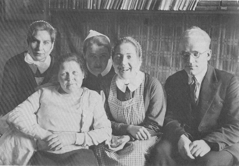 Mustavalkoinen kuva, jossa viisi henkilöä istuu lähellä toisiaan kirjahyllyn edessä, mukana kolme naista, yksi mies ja yksi nuorempi nainen, diakonissa.