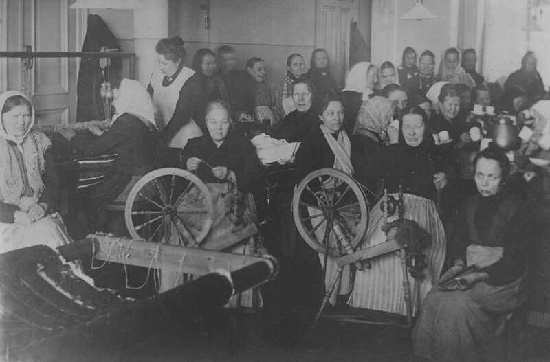 Mustavalkoinen historiallinen kuva, jossa ryhmä naisia istuu ja työskentelee käsityön parissa sisätiloissa, osa käyttää huiveja tai esiliinoja.