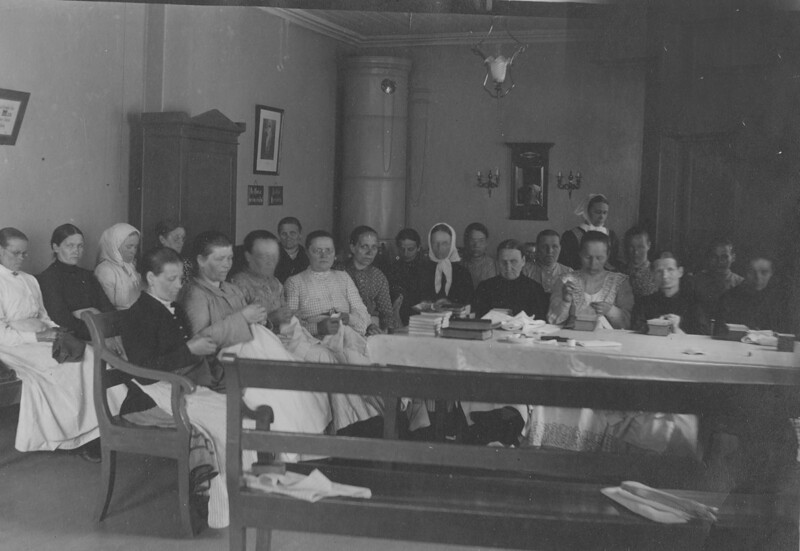 Mustavalkoinen kuva, jossa suuri ryhmä naisia istuu pitkän pöydän ympärillä sisätilassa, osa käsityövälineiden kanssa, taustalla huonekaluja ja koristeita.