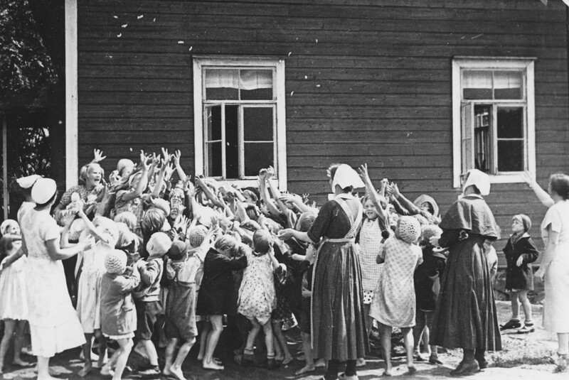 Mustavalkoinen valokuva, jossa lapset ja diakonissat leikkivät yhdessä ulkona vanhan puutalon edustalla.