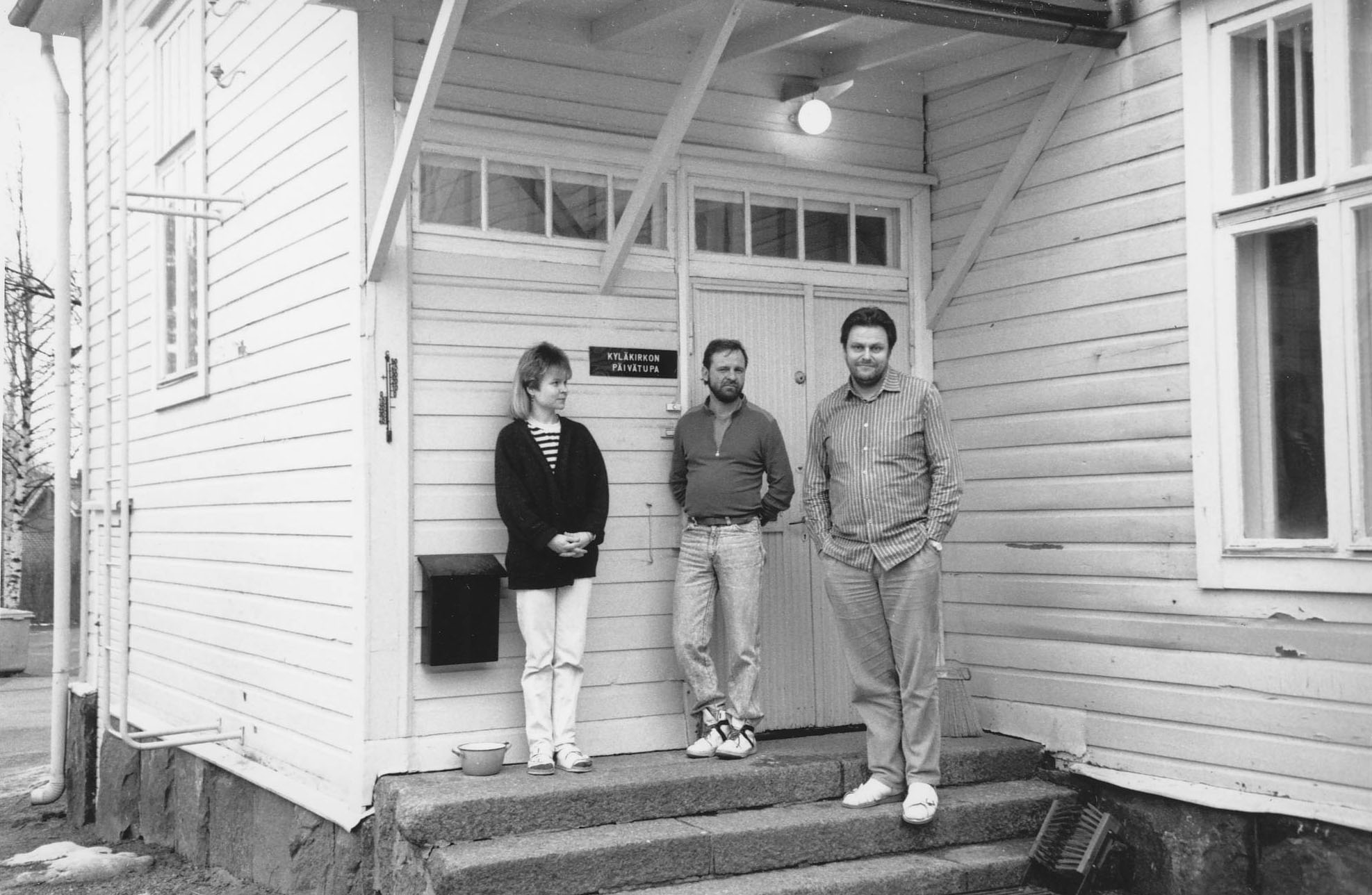 Kolme ihmistä seisoo puutalon portailla.