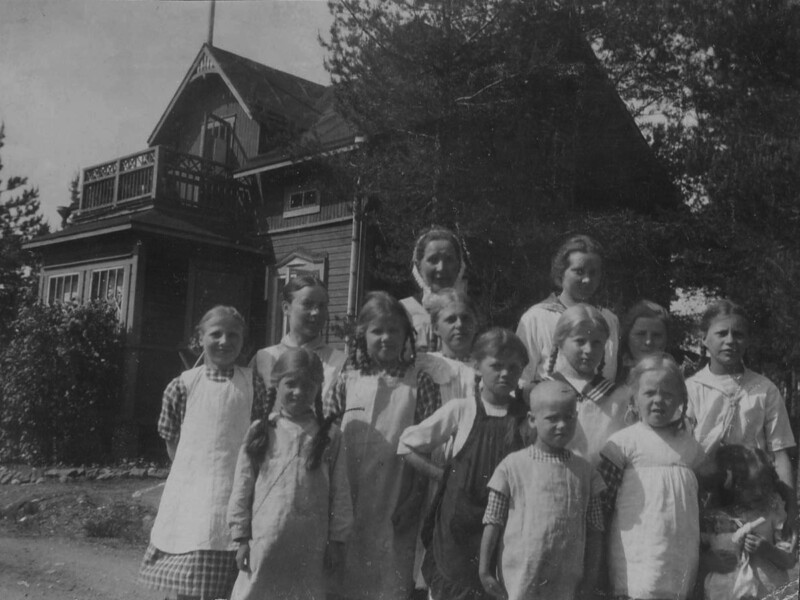 Mustavalkoinen valokuva, jossa ryhmä lapsia seisoo ulkona vanhan puutalon edustalla.