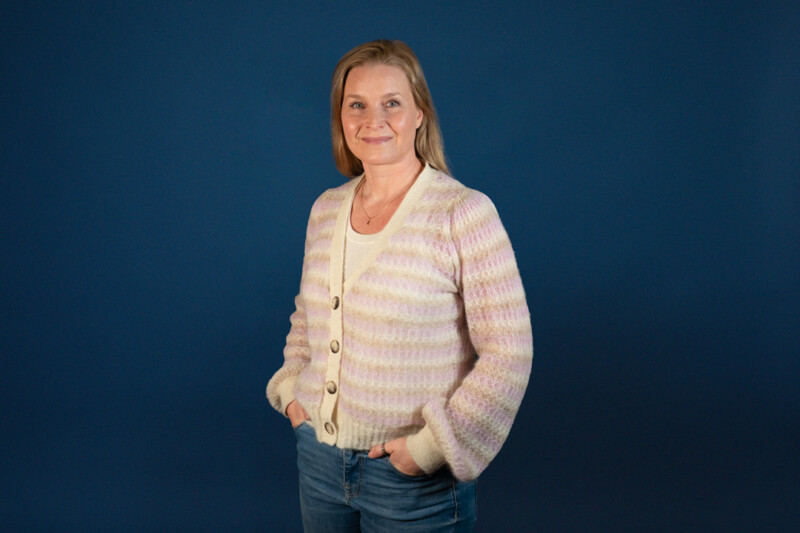 Vaaleaan neuletakkiin ja sinisiin farkkuihin pukeutunut vaaleahiuksinen nainen katsoo hymyillen kameraan.