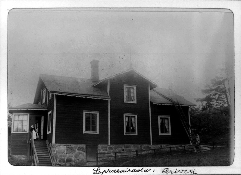 Mustavalkoinen valokuva vanhasta puutalosta, jossa on kiviperustus ja useita ikkunoita, talon edustalla kaksi henkilöä portailla.