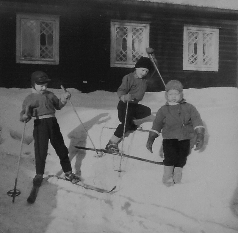 Kolme lasta leikkii lumessa talon edessä, kaksi heistä hiihtää ja yksi seisoo lumessa, mustavalkoinen kuva.