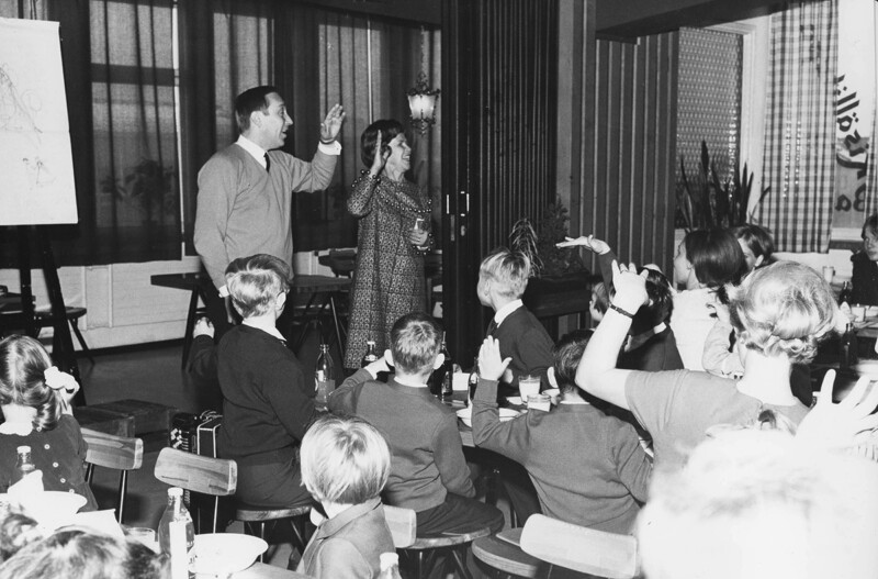 Mustavalkoinen kuva, jossa aikuinen mies ja nainen seisovat pöydän ääressä ja ohjaavat lapsia, jotka istuvat pöydän ympärillä nostamassa käsiään.