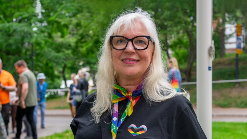 Vaaleahiuksinen nainen katsoo hymyillen kameraan. Hänellä on yllään paita, jonka rinnassa on sateenkaaren värinen sydän sekä kaulassaan sateenkaaren värinen kaulanauha.