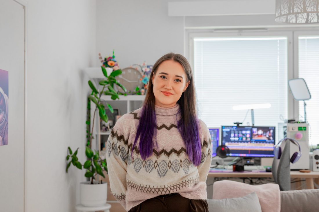 Yrityslahjoitus – Nuori nainen katsoo hymyillen kameraan. Taustalla asunto, jossa näkyy viherkasveja ja pöydällä tietokone.