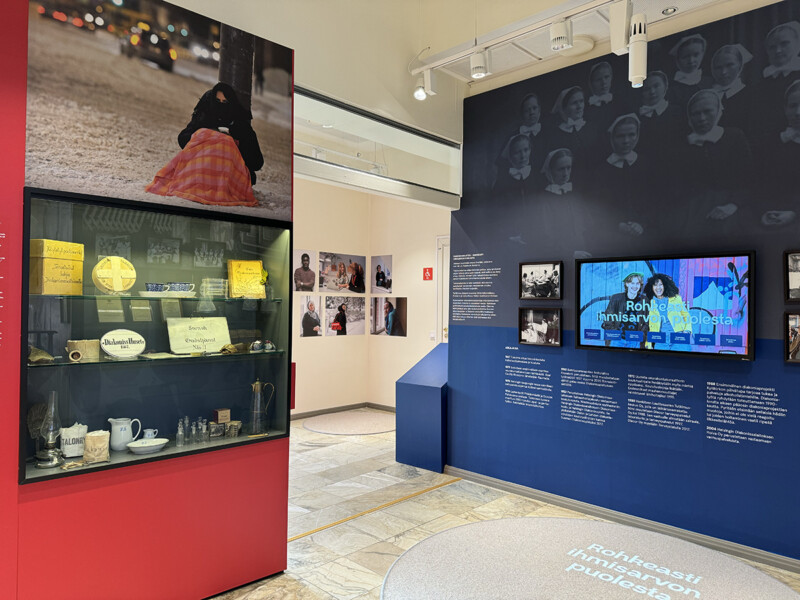 Kuva diakonissalaitoksen näyttelystä, jossa näkyy esinekaappi punaisella taustalla, seinälle ripustettuja valokuvia ja sinisellä seinällä näyttöruutu sekä tekstejä.