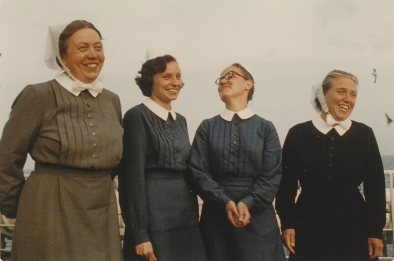 Neljä hymyilevää diakonissaa perinteisissä asuissaan ulkona, taustalla sininen taivas.