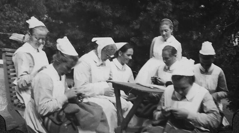 Mustavalkoinen kuva, jossa ryhmä naisia istuu yhdessä ulkona ja ompelee tai tekee käsitöitä, kaikki pukeutuneina perinteisiin hoitajavaatteisiin ja päähineisiin.
