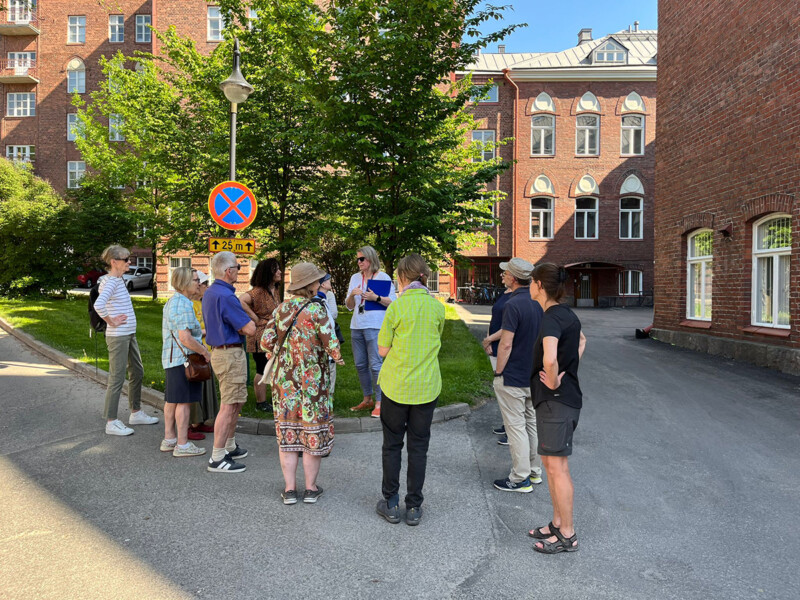 Opastus, jota ryhmä ihmisiä kuuntelemassa aurinkoisella kadulla, taustalla vanhoja tiilirakennuksia ja puita.
