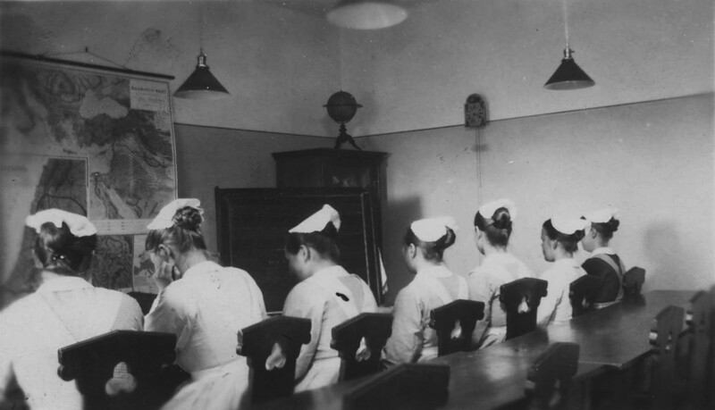 Mustavalkoinen kuva 1930-luvulta, jossa seitsemän diakonissalaitoksen oppilasta istuu rivissä selin kameraan luokkahuoneessa.