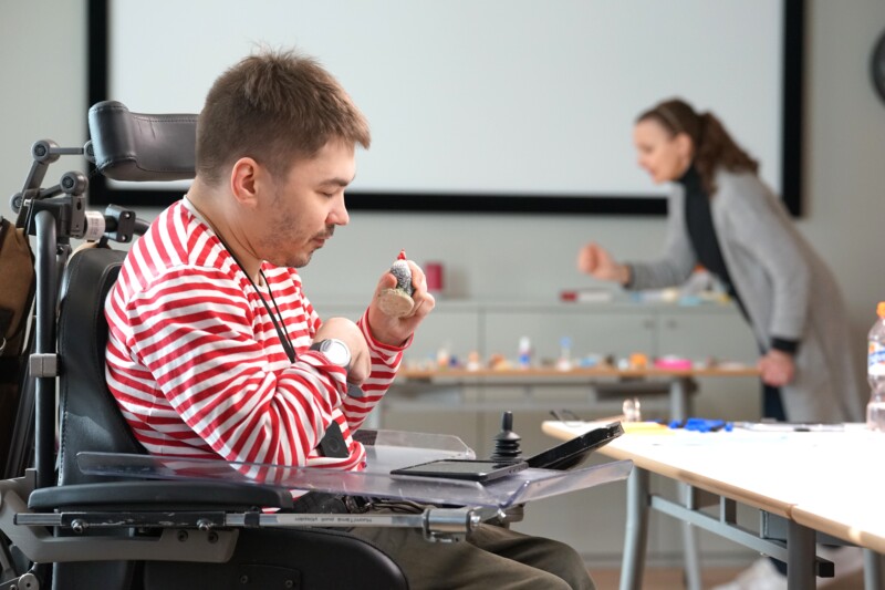 A young man in a wheelchair wearing a red and white striped shirt is engaging in an activity with small objects on a table, while a woman in the background is also participating in the activity in a bright room.