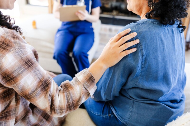 Kaksi ihmistä istuu rinnakkain. Toinen ihmisistä pitää kättään toisen henkilön olalla.