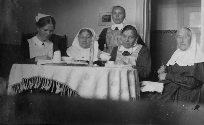 Mustavalkoinen kuva, jossa viisi naista istuu pöydän ympärillä sisätiloissa; naiset pukeutuneina diakonissalaitoksen asuihin ja pääsuojiin.