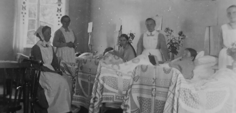 Mustavalkoinen historiallinen valokuva, jossa sairaanhoitajia ja potilaita sairaalasalissa.