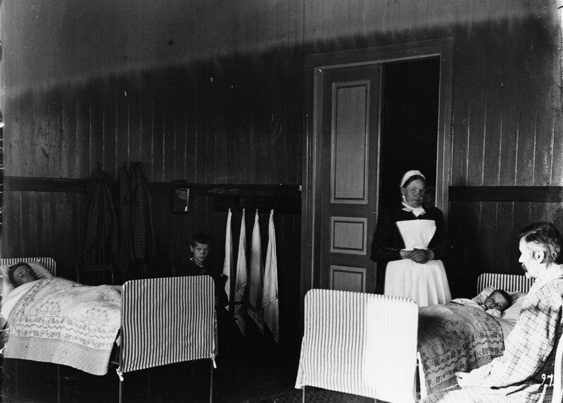 Mustavalkoinen kuva sairaalaympäristöstä, jossa kaksi potilasta makaa vuoteissaan ja diakonissa seisoo oviaukossa valkoisessa essussa ja päähineessä.