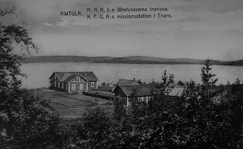Mustavalkoinen valokuva, jossa näkyy kaksi puurakennusta järven rannalla metsäisessä maisemassa, teksti: "RIUTULA. N.N.K.Y:n lähetysasema Inarissa. K.F.U.K:s missionsstation i Enare."