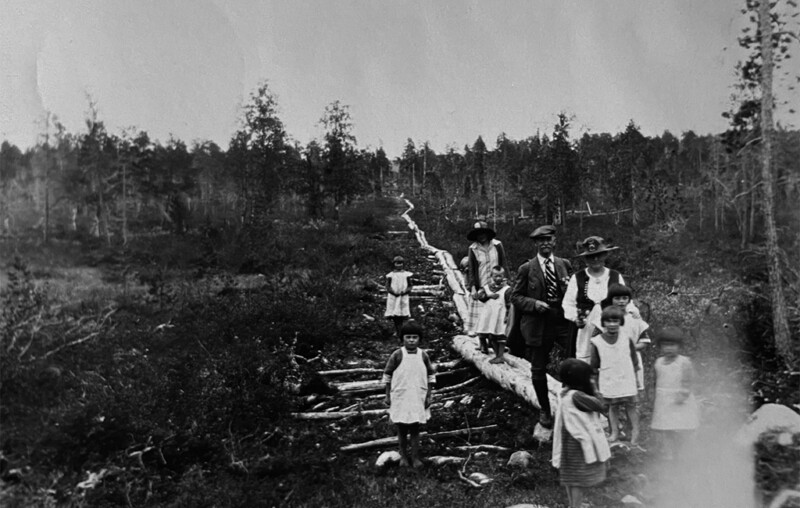 Mustavalkoinen historiallinen valokuva, jossa ryhmä ihmisiä, mukaan lukien aikuisia ja lapsia, kävelee pitkospuilla metsäisessä maastossa.