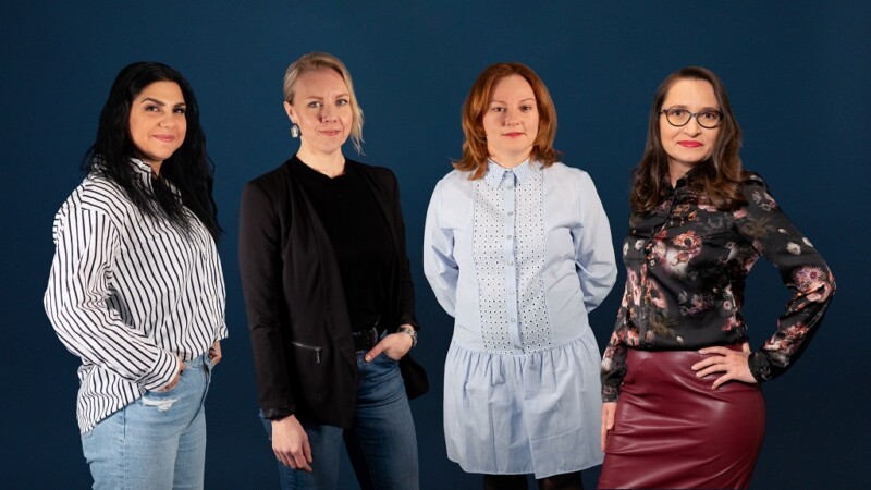 Four women standing side by side against a dark blue background, dressed in casual and semi-formal attire, looking at the camera with confident expressions.
