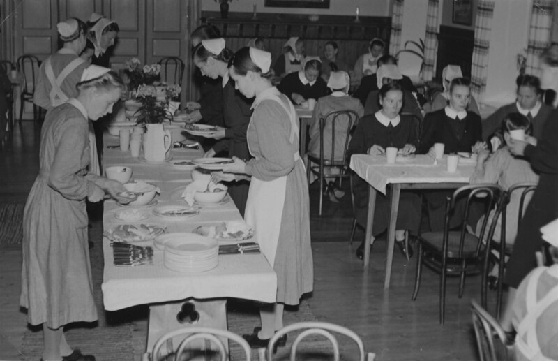 Mustavalkoinen kuva 1950-luvun diakonissalaitoksen ruokasalista, jossa naisia ja tyttöjä tarjoilemassa ja istumassa pöytien ääressä.