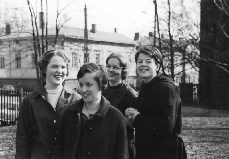 Mustavalkoinen valokuva, jossa neljä nuorta naista seisoo ulkona puiden ja rakennuksen edessä 1960-luvulla, kaikki hymyilevät ja katsovat eri suuntiin.