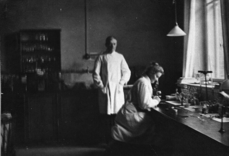 Mustavalkoinen valokuva 1900-luvun alun tutkimuslaboratoriosta, jossa yksi henkilö seisoo ja toinen istuu työpöydän ääressä tutkimassa näytteitä.