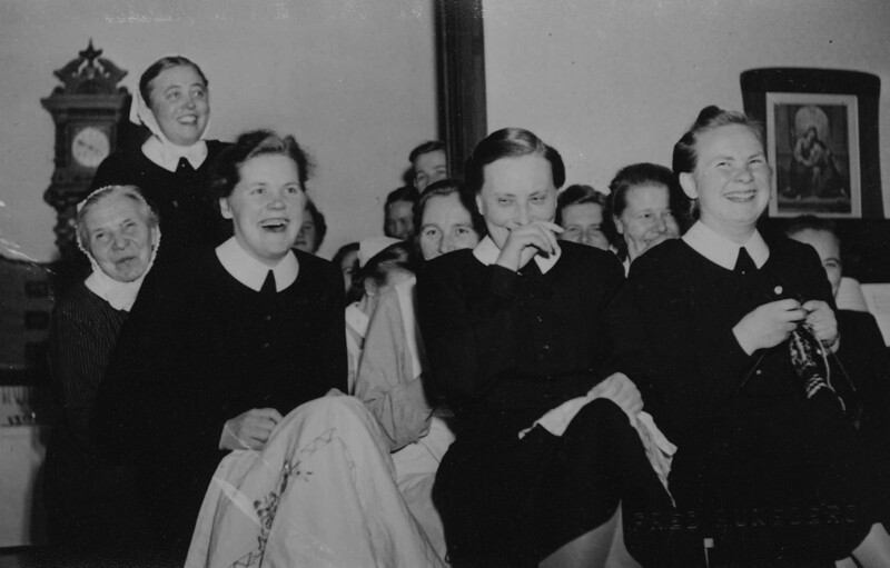 Mustavalkoinen kuva, jossa ryhmä naisia istuu vierekkäin huoneessa ja hymyilee tai nauraa.