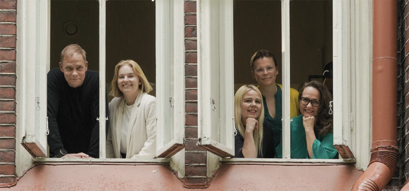 Neljä naista ja yksi mies katsovat ulos kahdesta vierekkäisestä ikkunasta vanhassa rakennuksessa, hymyillen ystävällisesti.
