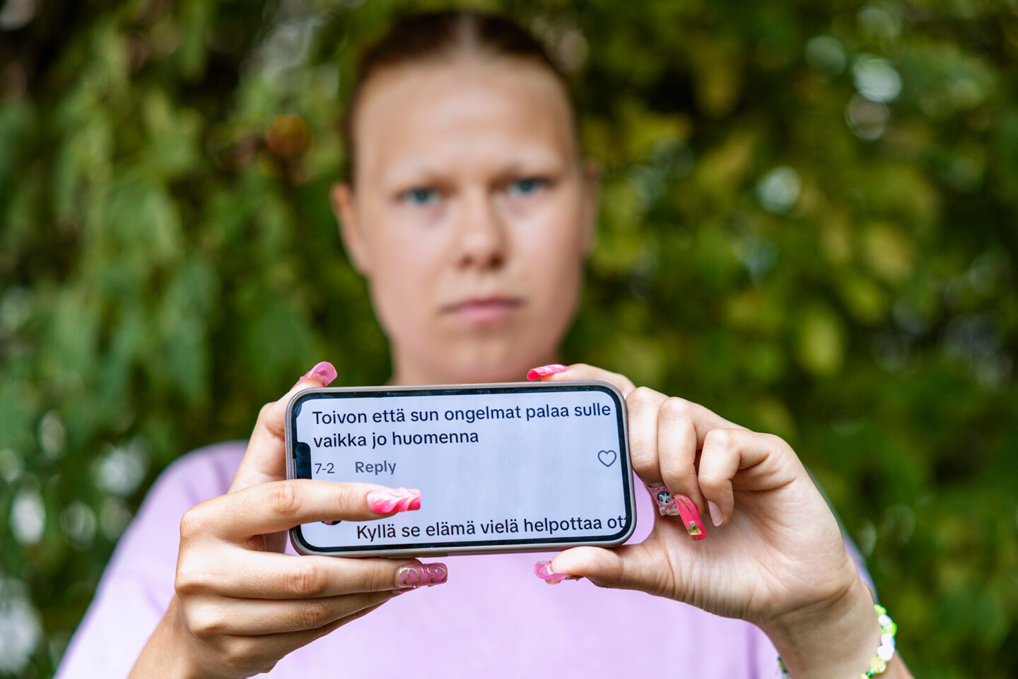 Henkilö pitelee kädessään puhelinta, jossa näkyy somekommentteja, taustalla epäselvästi vihreää kasvillisuutta.