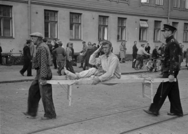 Mustavalkoinen historiallinen valokuva, jossa kaksi miestä kantaa potilasta kantotuolissa kadulla, taustalla ihmisiä ja vanhoja rakennuksia.