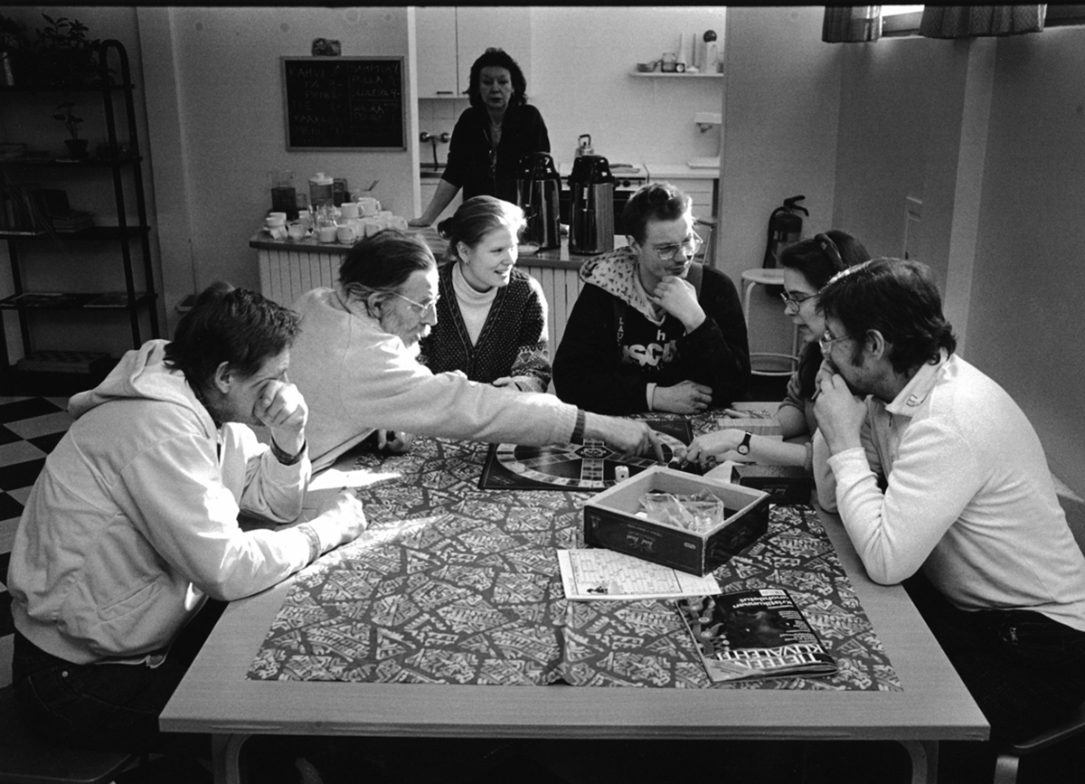 Mustavalkoinen kuva, jossa ryhmä ihmisiä istuu pöydän ympärillä pelaamassa lautapeliä, taustalla nainen seisoo kahvipöydän luona.